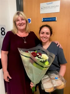 a patient and dentist pose for a photograph with flowers and baked goods